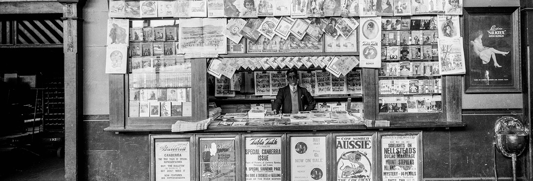 Newspaper stand at Spencer St train station, July 1930, VPRS 12903/P1 Box 23 Item 1 Black and white image of man working at newspaper stand, Spencer St train station