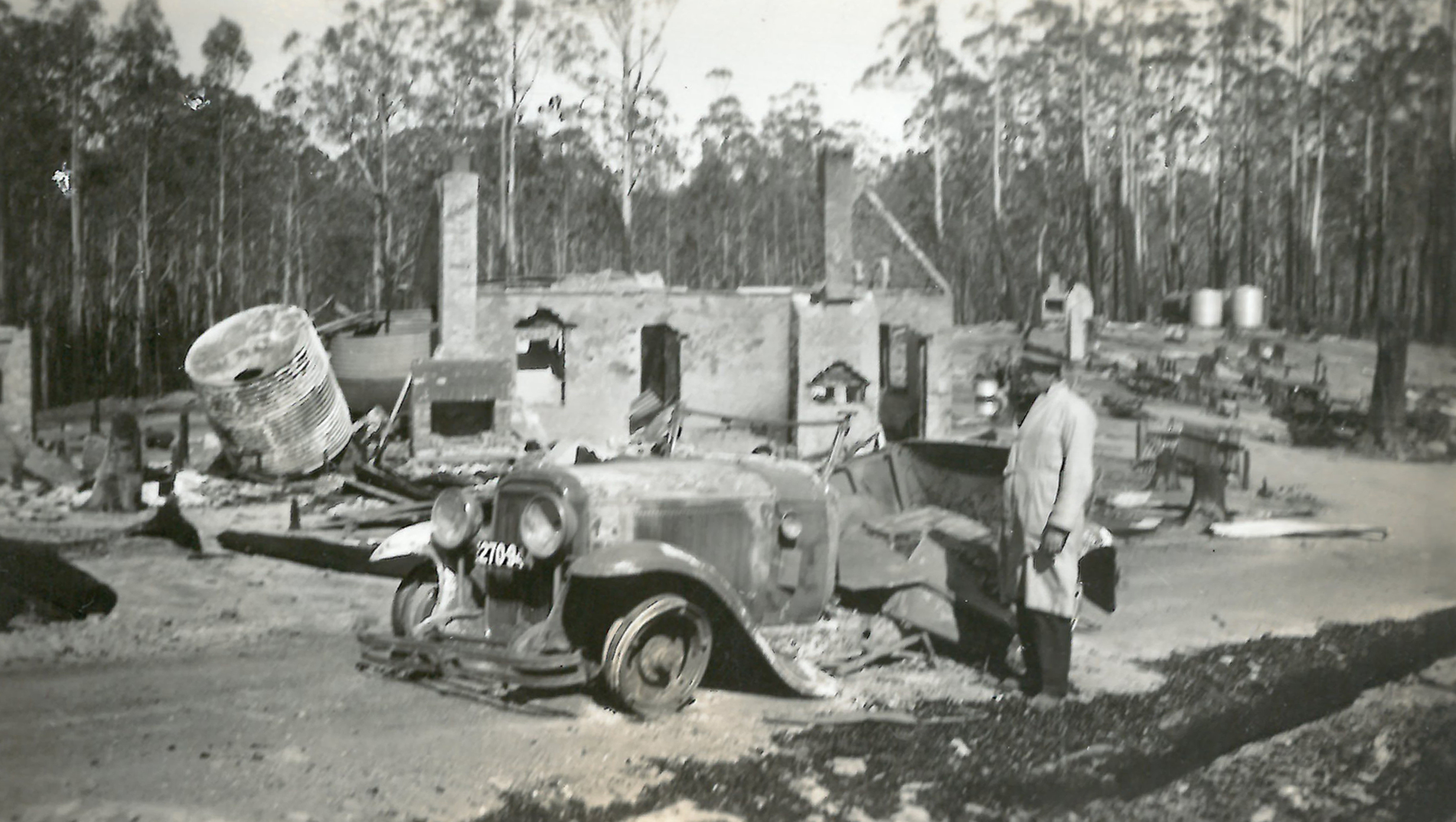 black and white photo of burned hut and car with man standing in front